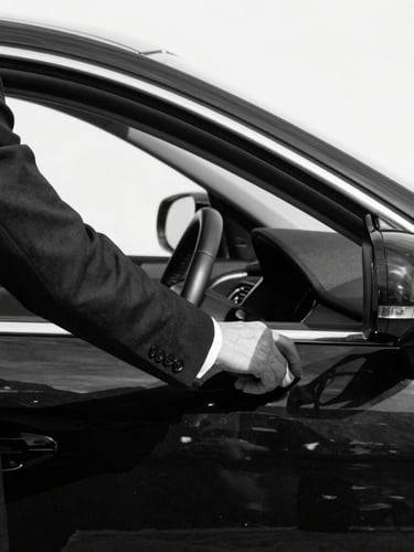 Tight macro shot of a driver in a sharp dark suit opening a polished black sedan door, high-contrast natural light, sharp shadows, desaturated black and white tones