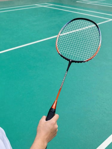Candid daylight shot of a young girl's hands holding a badminton racket, details of the grip, with bright teal court surface in the background.