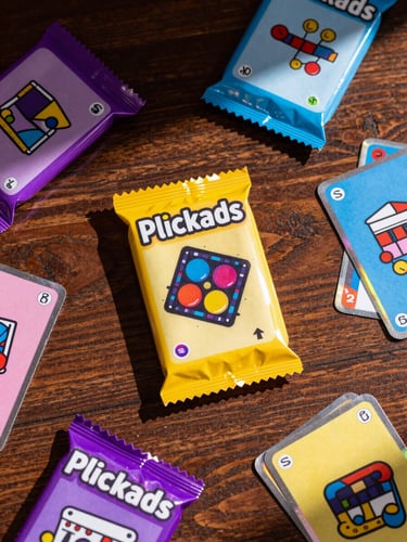 Overhead flat-lay of colorful Plickcards booster packs and loose cards scattered across a dark wooden school desk, sharp dramatic shadows and bright highlights.