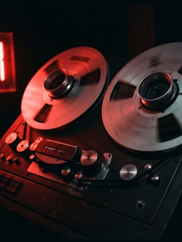 Cinematic close-up of analog tape reels spinning in a dark studio, harsh red neon light reflecting on metallic surfaces, heavy 35mm film grain, moody chiaroscuro shadows.