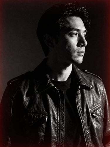 High-contrast black and white portrait of an artist in a leather jacket, looking away, dark shadows, warm tungsten backlight, subtle grain, artistic film aesthetic.