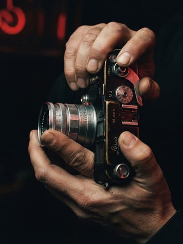 Cinematic close-up of a vintage 35mm film camera held by hands, warm tungsten light, heavy analog film grain, dark studio background, red neon reflection.