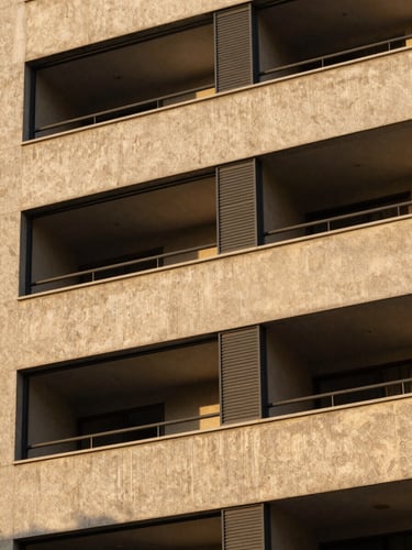 Close-up architectural detail of a completed residential building facade, warm golden-hour light raking across textured concrete and dark metal window frames, sharp material depth, a resident visible through glass in an upper floor, Brazilian urban context
