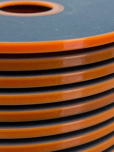 Macro shot of freshly cured polyurethane sealing discs stacked in an Indonesian engineering facility, crisp high-contrast lighting highlighting the glossy orange elastomer edges.