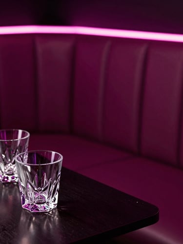 Low-light close-up of a plush leather VIP booth illuminated by soft magenta neon accents, pristine crystal glassware on a dark table, high contrast.