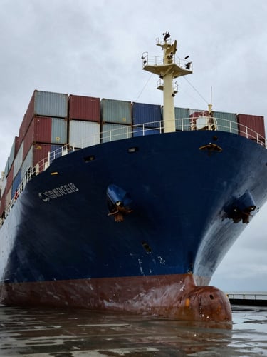 Low-angle shot of a massive container ship hull in a drydock, dramatic overcast sky, towering steel structure, wet concrete ground, industrial maritime photography