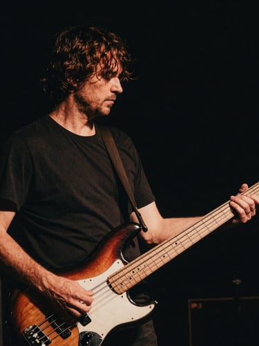 Moody low-key photography of Pierre-Yves Clarence holding his electric bass, high contrast stage lighting, deep shadows, warm amber undertones, 35mm film grain.
