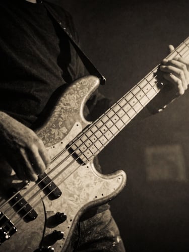 Close-up of bassist hands on the strings of an electric bass, dramatic side lighting, high contrast monochrome, warm amber undertones, smoky jazz club atmosphere.