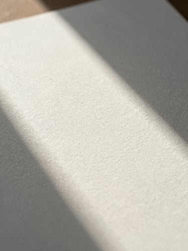 Extreme close-up of premium textured watercolor paper with soft morning shadows casting a clean geometric line across the surface.