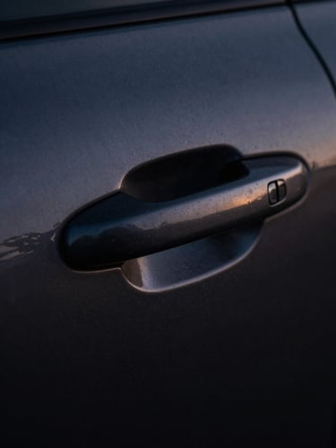 Extreme close-up of a dark brushed metal vehicle door handle, dramatic single-source shadow, cool twilight ambient light, high-contrast.