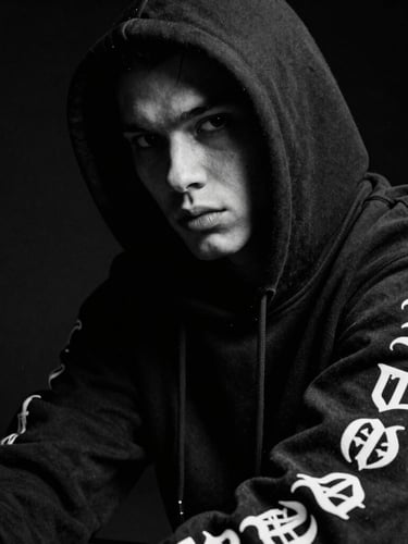 High-contrast black and white close-up of a rebellious model wearing a heavy black hoodie with gothic lettering on the sleeve, dramatic studio lighting, dark background, 35mm film grain.