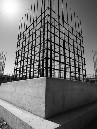Low-angle architectural shot of a reinforced concrete foundation footing on an active construction site in Bareilly, steel rebar grid visible, harsh midday sun, high-contrast concrete tones