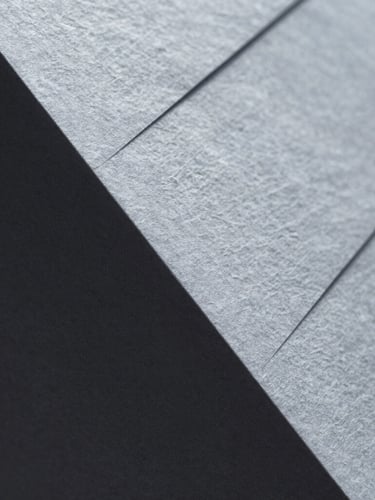 Macro close-up of high-texture heavy cotton paper with blind debossed geometric lines, sharp side-lighting, silver-gray shadows, minimalist architectural feel.