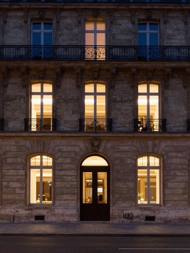 Symmetrical wide shot of a classic Parisian stone building facade with warm interior window lights at dusk, clean architectural lines, deep shadows, 35mm lens