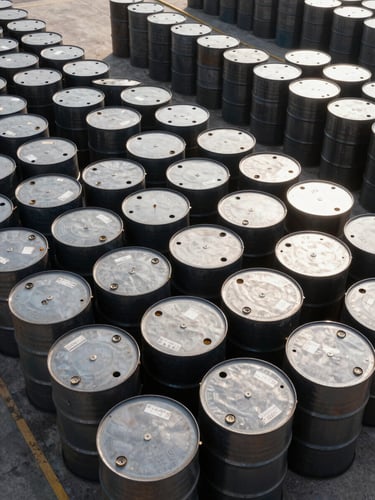 Overhead view of stacked steel crude oil barrels at a port terminal, daylight, grade and origin markings visible on barrel heads, no people, industrial concrete surface, tight framing showing rows receding into distance