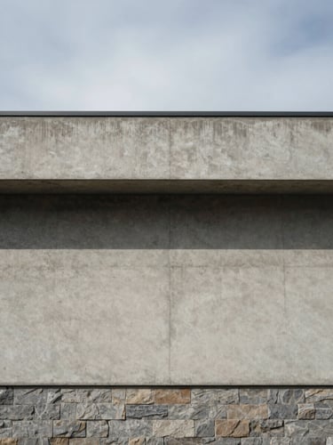 Eye-level exterior view of a raw concrete facade under clear overcast daylight, a deep horizontal shadow band marking the window reveal, muted stone cladding visible at the base, wide environmental framing with open sky above