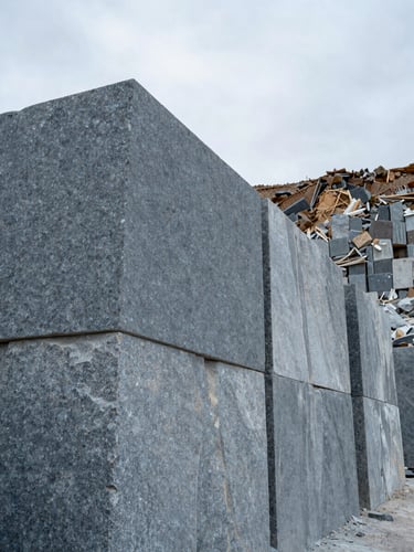 Wide-angle close view of stacked granite blocks in a Bolivian quarry, natural overcast daylight illuminating the grey-blue stone surface texture, quarry wall and open sky visible in the background right third, generous empty sky on the upper left