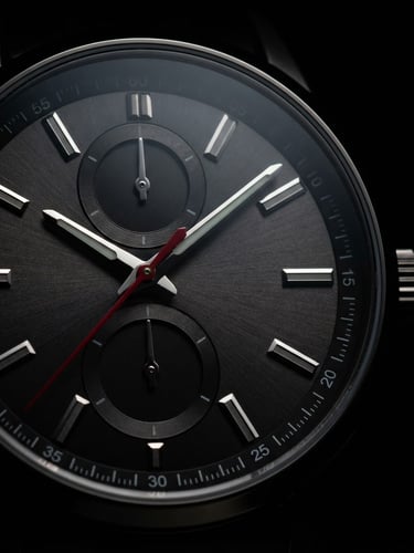 Macro close-up of a MaimAshi watch dial, dramatic low-key studio lighting with sharp rim lights highlighting the metallic edges and crimson second hand against pure black.
