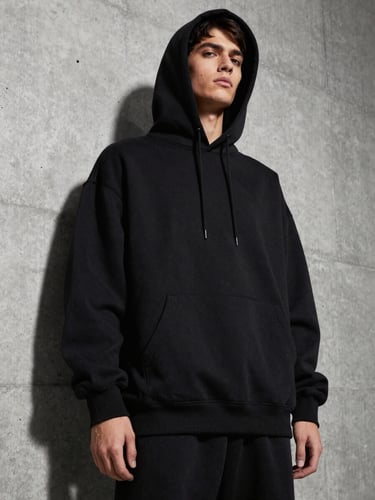 Low-angle medium shot of a model wearing a heavy black oversized hoodie, standing against a stark concrete wall, dramatic side shadows highlighting the dense fabric texture.