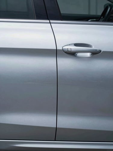 Split-frame straight-on documentation of a car door panel: left half shows a sharp horizontal dent crease, right half shows the same surface after PDR repair — perfectly smooth — bright shadowless studio light, cold steel metallic paint
