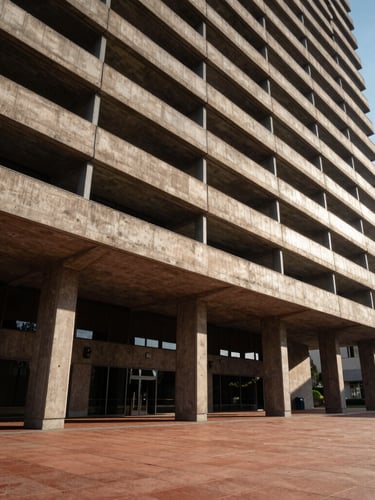 Low-angle architectural shot of a brutalist concrete and steel commercial tower facade meeting a warm terracotta-tiled plaza under high-contrast afternoon sun, casting deep geometric shadows.