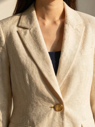 A close-up portrait of a woman wearing a tailored linen blazer in soft cream, warm golden-hour window light casting soft shadows, focus on the delicate stitching and gold button details