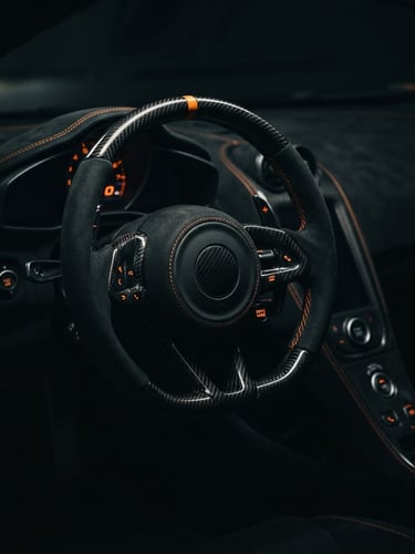 Low-angle dramatic close-up of a high-performance sports car steering wheel, carbon fiber detailing, sharp orange stitching, moody ambient lighting