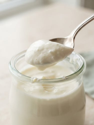 Macro close-up of thick, velvety yogurt on a silver spoon, elegant glass jar on a clean stone countertop, soft natural morning daylight, minimalist cream and sage tones