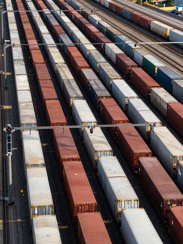 Panoramic overhead view of a freight rail classification yard, rows of intermodal container cars sorted by corridor, track switches and signal gantries visible, full daylight, strong diagonal geometry filling the frame edge to edge