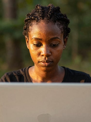 A close-up shot of a young Nigerian woman focusing on a laptop screen, warm golden hour sunlight illuminating her face, shallow depth of field, professional documentary style.