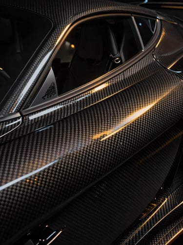 Tight macro shot of polished carbon fiber weave on a modern supercar body, dramatic low-key studio lighting with sharp gold reflections, deep shadows, high contrast, 50mm