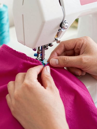 Close-up of hands carefully stitching a vibrant magenta pet garment on a sewing machine, warm natural daylight, spool of teal thread visible