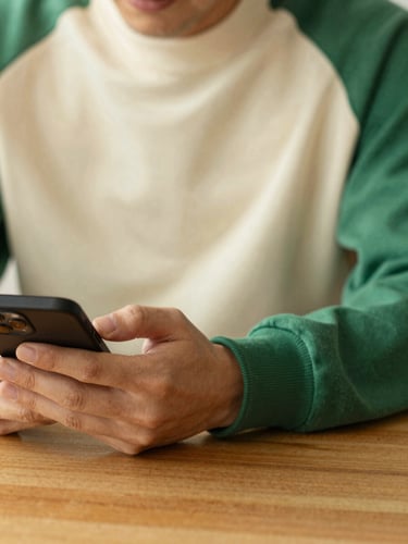 Warm editorial portrait of a person sitting at a wooden table with a phone, soft natural daylight, shallow depth of field, rich cream and green tones.