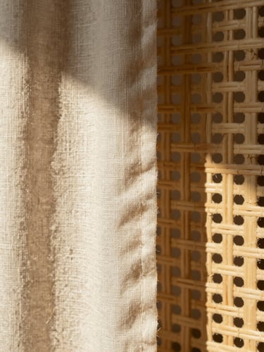 Macro shot of raw unbleached linen texture beside a hand-woven rattan screen, dramatic morning light casting high-contrast shadows, warm sandy-beige tones, 35mm lens
