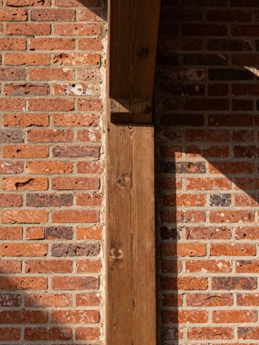 Straight-on architectural detail crop of a weathered red brick wall meeting a massive hand-hewn oak support beam, warm natural daylight