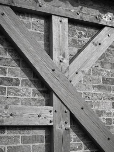 Black and white architectural detail crop of heavy timber framing and original iron bolts in a brick wall, natural side lighting, high contrast.