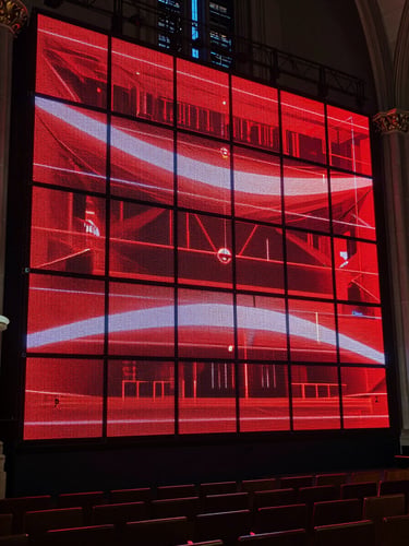 Wide environmental shot of a large installed LED wall dominating the front of a darkened church sanctuary, the screen blazing with deep red and white graphics during a live production, cinematic tungsten stage lighting raking across the room from the sides, rows of empty seats in the foreground, the wall's seamless panels glowing with controlled precision