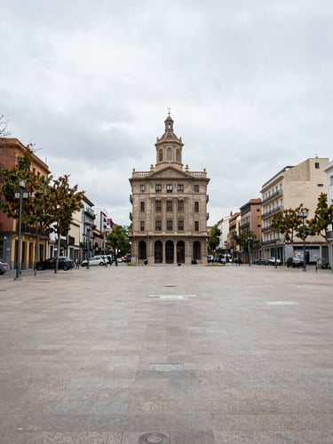 Wide-angle honest documentary photo of an empty public square in Catalonia in natural overcast daylight, 35mm lens, realistic street-level composition.