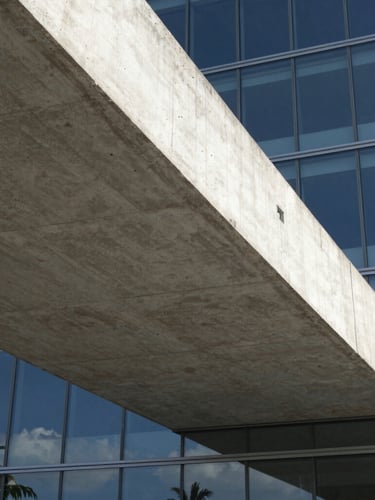 Extreme low-angle view of a dramatic concrete cantilever slab meeting a glass facade, sharp geometric shadows, bright coastal daylight
