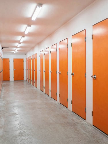 Wide-angle interior shot of a brightly-lit, clinical self-storage corridor in Al Quoz, clean concrete floor, orange unit doors, overhead LED lighting, sharp perspective.