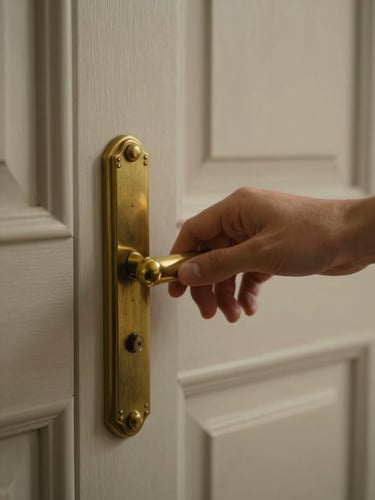 Close detail of a hand resting on a door handle, aged brass hardware on a painted wooden door, soft diffused light, muted warm background, no face visible