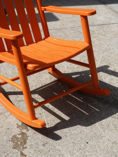 A single orange resin rocking chair casting a sharp, graphic shadow on a wet concrete patio under direct sunlight, texture-focused close-up