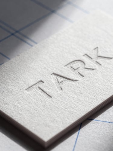 Stark macro photography of a physical embossed business card on heavy cotton paper, sharp directional studio light casting deep shadows, technical blue grid lines faintly visible.