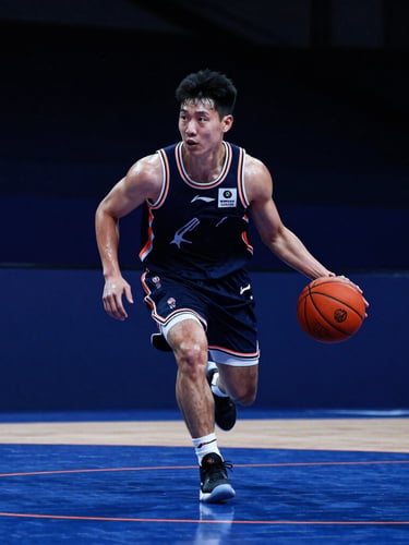 Dynamic low-angle shot of a basketball player mid-dribble on an indoor court, dramatic side-lit sweat, high-contrast stadium lighting, deep blue atmosphere with sharp orange court lines