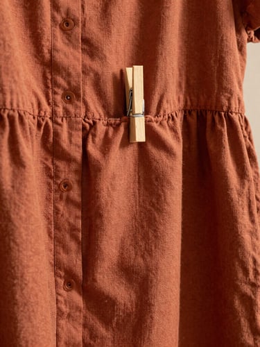 Close-up of a pattern-drafted linen-blend dress hanging on a raw wooden peg, warm clay terracotta fabric texture, soft side-lighting.