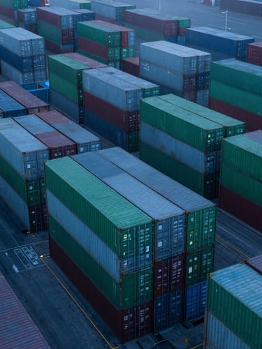 High-altitude drone photograph of a geometric container terminal at twilight, cool blue tones, perfectly aligned stacks of green and grey shipping containers, misty atmosphere.