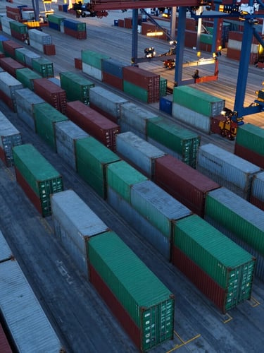 An overhead drone shot of a massive container terminal at dusk, cool twilight lighting, sharp geometric rows of green and grey shipping containers, clean composition.