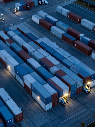 High-altitude drone shot of a modern geometric container terminal at twilight, cool blue lighting, structured rows of cargo
