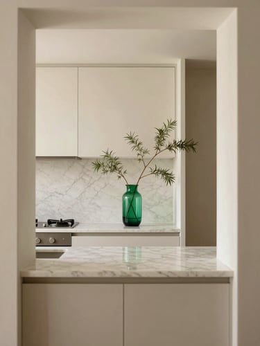 Straight-on architectural photograph of a minimalist kitchen counter, warm natural light, pristine marble surfaces, emerald glass vase with single branch, 35mm