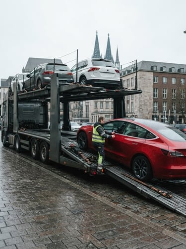 Low-angle wide shot of a professional transport trailer loading a modern sedan on a clean Cologne street, cool overcast daylight, desaturated tones with vibrant red details, 35mm lens